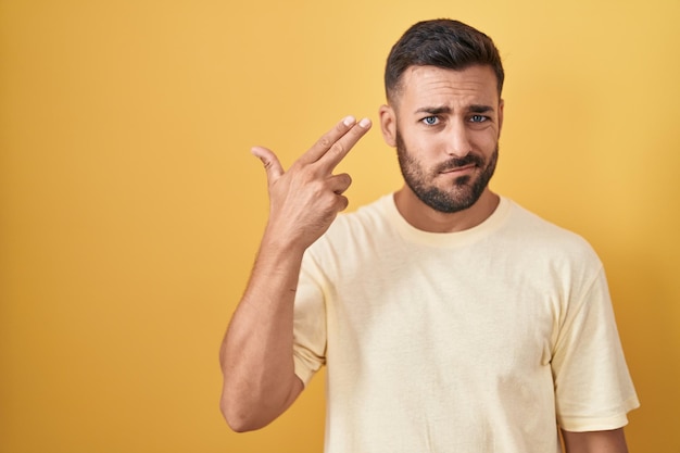 Handsome hispanic man standing over yellow background shooting and killing oneself pointing hand and fingers to head like gun, suicide gesture.