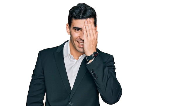 Handsome hispanic man wearing business clothes covering one eye with hand, confident smile on face and surprise emotion.