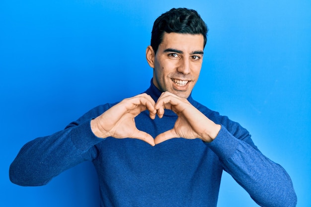 Handsome hispanic man wearing turtleneck sweater smiling in love doing heart symbol shape with hands romantic concept