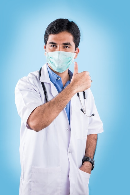 Handsome Indian Male Doctor with surgical or safety health Mask on. standing isolated over blue background