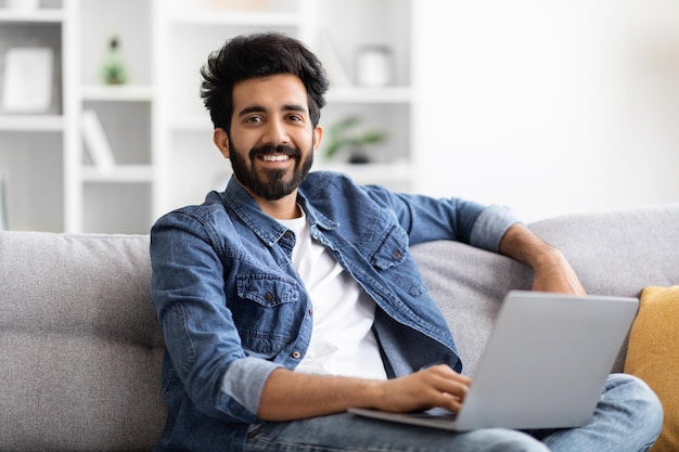 Handsome indian male using laptop while sitting on couch at home