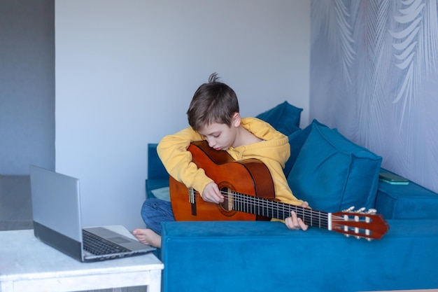 Handsome kid dressed casual jeans and yellow sweatshirt watches video lesson on playing the guitar on the cozy sofa at home