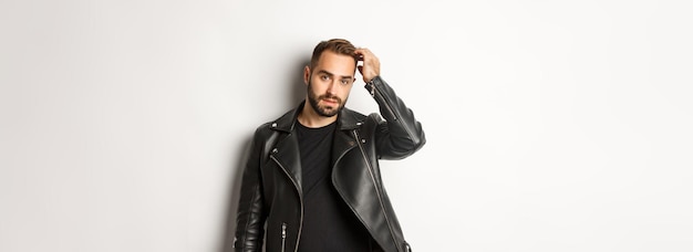 Handsome macho man in black biker jacket touching his haircut and looking cool white background