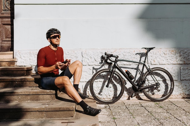 Handsome male cyclist wearing sport clothes surfing internet on smartphone while resting outdoors Beautiful summer weather outdoors