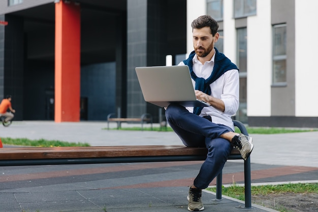 Handsome male freelancer in stylish clothing sitting on wooden bench and using modern laptop. Bearded man working outdoors with portable computer.