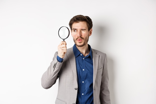 Handsome male model in suit looking through magnifying glass with interest, seeing something aside