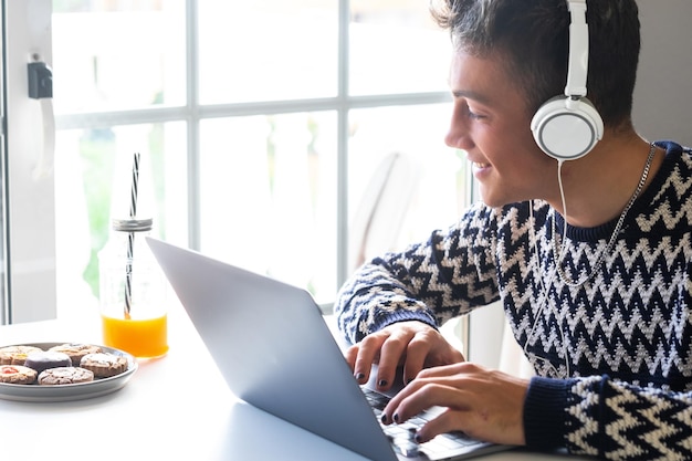 Handsome male teenager using laptop at home following an online lesson White desk with food and drink