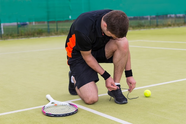 Handsome male tennis player tying shoelaces wearing a sportswear left racket and ball on a green grass court outdoor in summer or spring