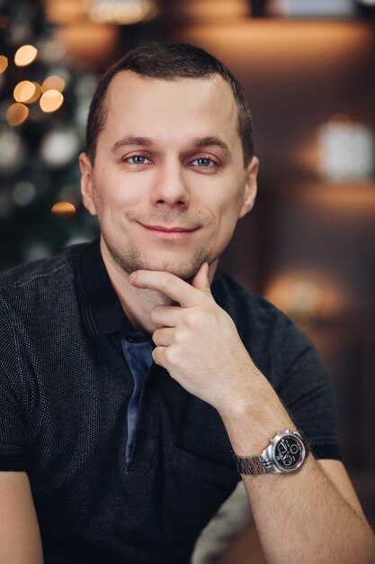 Handsome man against illuminated Christmas tree fashionable watch looking away with hand on chin and pensive look 