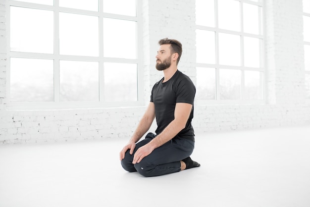 Handsome man in the black sportswear doing yoga in the white gym interior