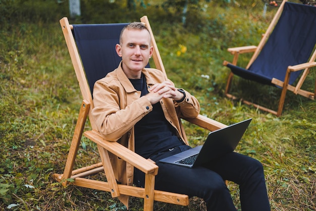 A handsome man in a brown shirt sits on a wooden folding chair in the garden and works with a laptop Wooden ecological furniture Remote work