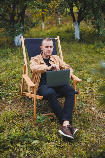 A handsome man in a brown shirt sits on a wooden folding chair in the garden and works with a laptop Wooden ecological furniture Remote work