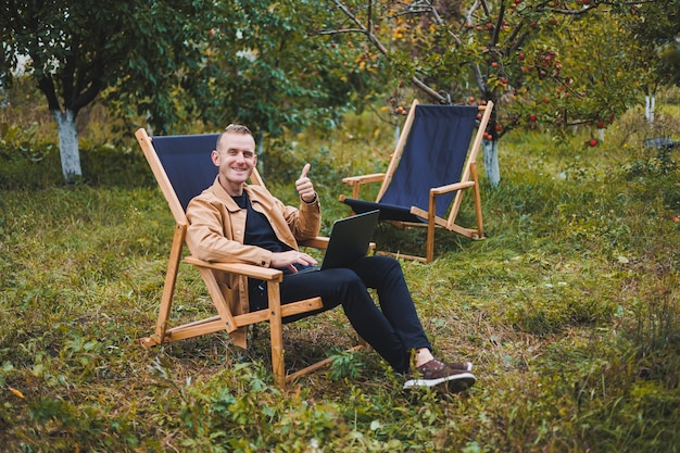 A handsome man in a brown shirt sits on a wooden folding chair in the garden and works with a laptop Wooden ecological furniture Remote work