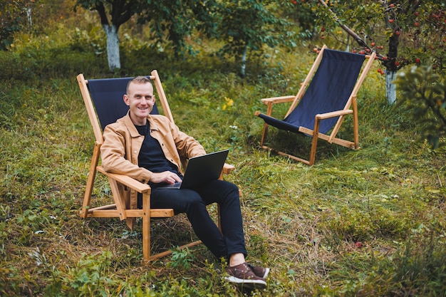 A handsome man in a brown shirt sits on a wooden folding chair in the garden and works with a laptop Wooden ecological furniture Remote work