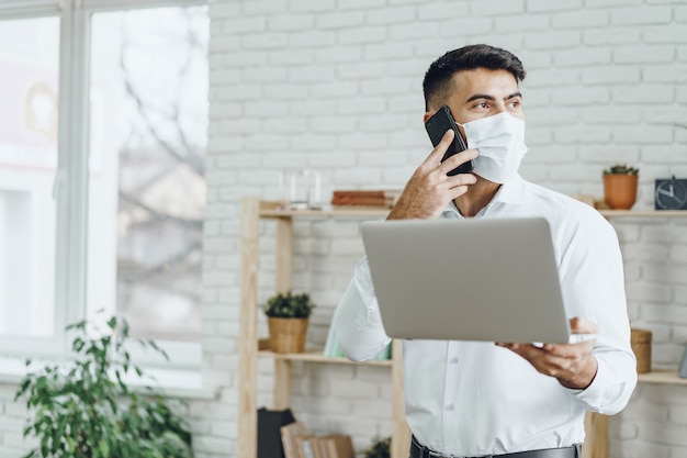 Handsome man businessman in medical mask using his laptop while talking on his cellphone indoors