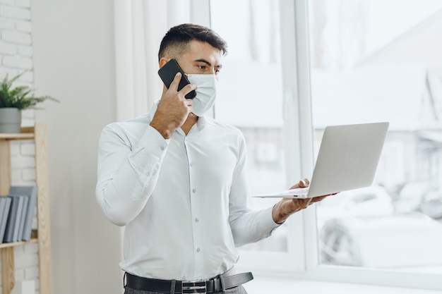 Handsome man businessman in medical mask using his laptop while talking on his cellphone