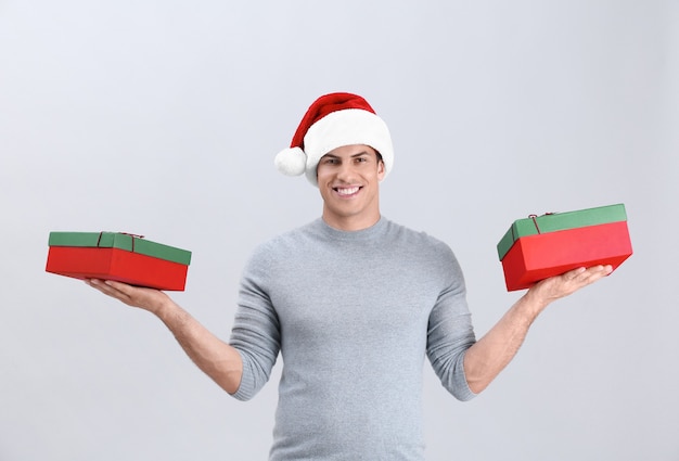 Handsome man in Christmas hat holding gift boxes on light background