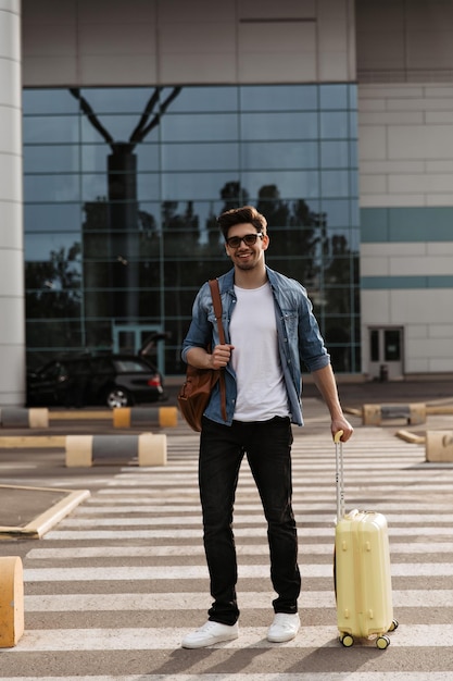 Handsome man in denim jacket white tshirt and black pants smiles Tourist in sunglasses holds yellow suitcase and brown backpack