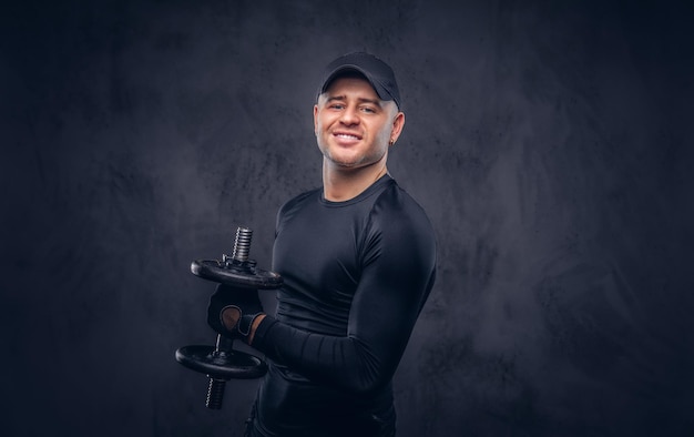 A handsome man, dressed in a black sportswear and baseball cap lifts a dumbbell.