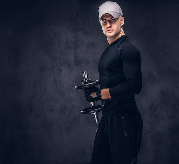 A handsome man, dressed in a black sportswear and baseball cap lifts a dumbbell.