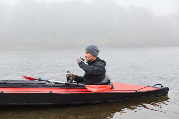 Handsome man drinking tea from thermos and warming while sitting in canoe in middle of river