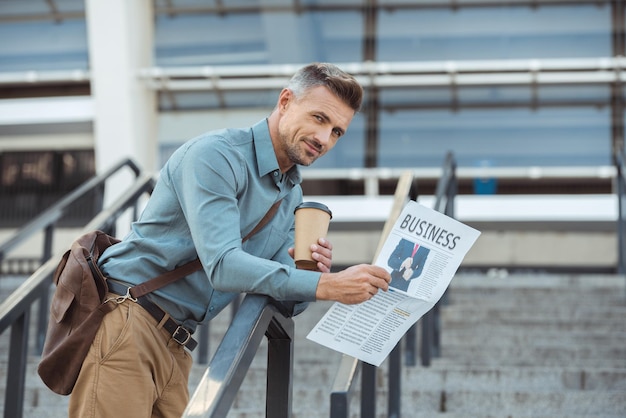 Handsome man holding business newspaper and coffee to go while standing on stairs and smiling at