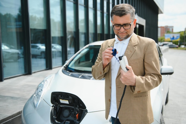 Handsome man holding charging cable at electric charging station point standing near his new car