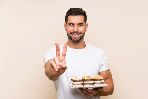 Handsome man holding muffin cake over isolated  smiling and showing victory sign