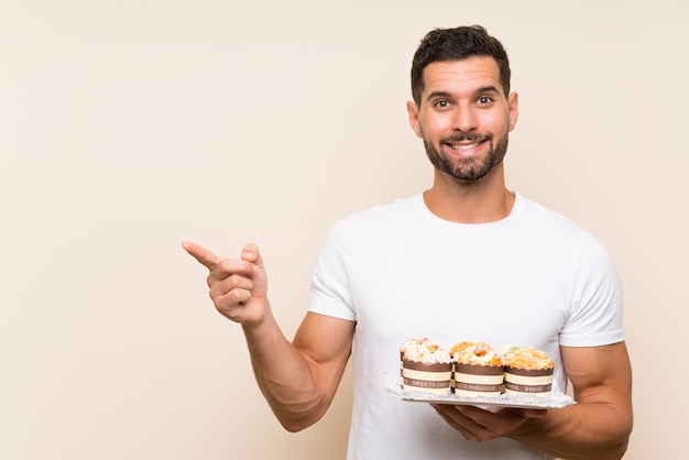 Handsome man holding muffin cake over isolated wall surprised and pointing finger to the side