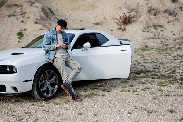 Handsome man in jeans jacket and cap is standing near his white muscle car in career and texting on phone