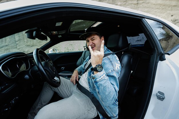 Handsome man in jeans jacket and cap sit at his white muscle car and show Rock and Roll hand sign