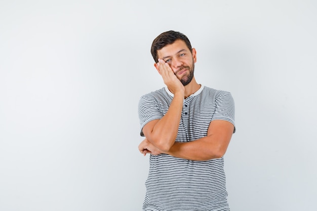 Handsome man keeping hand on cheek in t-shirt and looking fatigued , front view.