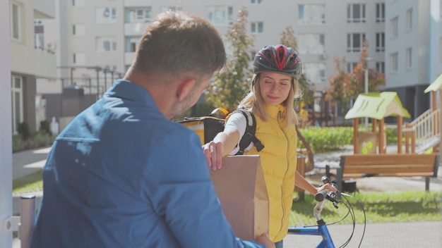 Handsome man open door to a food delivery woman wearing yellow thermal backpack on a bike and recieves order or package with food Food delivery concept