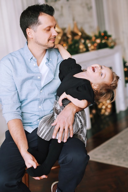 A handsome man plays with his funny daughter near the Christmas tree at home