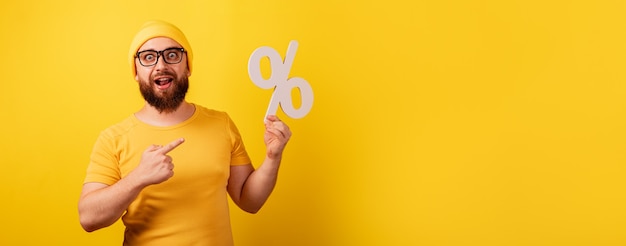 Handsome man pointing at percent symbol over yellow background, panoramic image with space for text