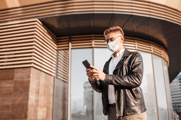 Handsome man in a protective mask on his face with glasses with a smartphone on the street of a big city Businessman talking on the phone on urban background