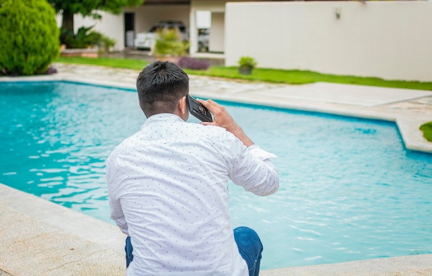 Handsome man sitting calling on the phone near the swimming pool Young man sitting calling on the phone by a swimming pool Guy sitting talking on the cell phone near the edge of the swimming pool