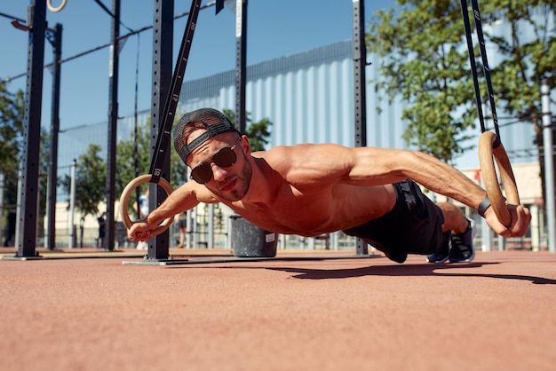 Handsome man on sport ground The sportsman the guy carries out difficult exercise sports gymnastics Muscular man doing workout with rings The concept of a modern young man