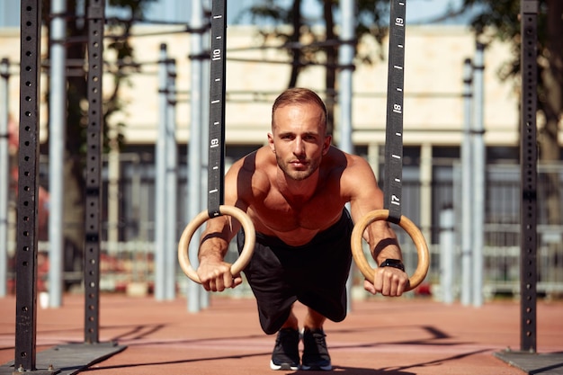 Handsome man on sport ground The sportsman the guy carries out difficult exercise sports gymnastics Muscular man doing workout with rings The concept of a modern young man