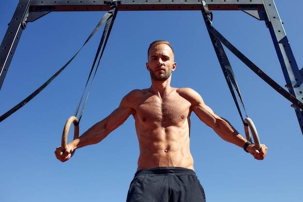 Handsome man on sport ground The sportsman the guy carries out difficult exercise sports gymnastics Muscular man doing workout with rings The concept of a modern young man