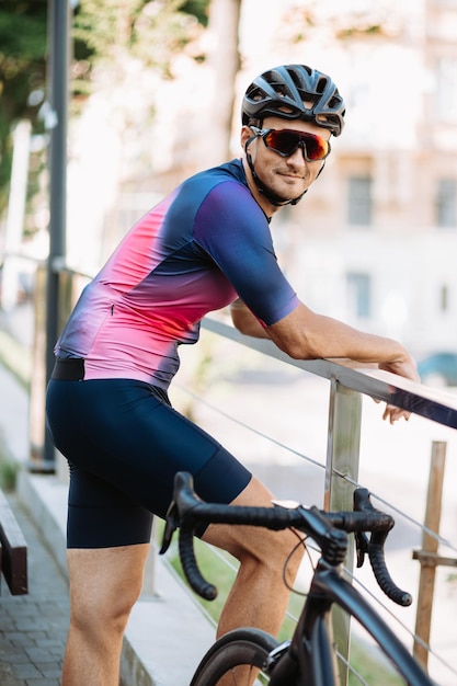 Handsome man in sportswear helmet and glasses smiling and looking at camera while standing near black bike outdoors Healthy lifestyles concept