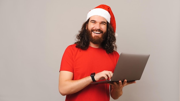 Handsome man wearing Christmas hat is holding a laptop while wide smiling