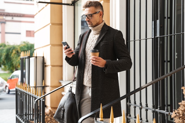 Handsome man wearing a coat walking outdoors, using mobile phone, drinking coffee