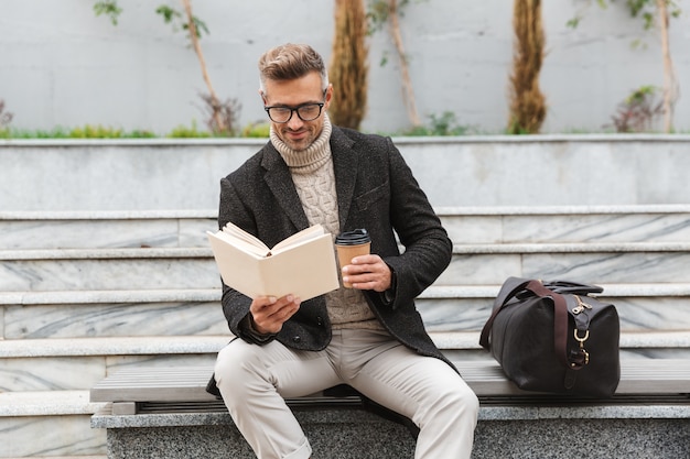 Handsome man wearing jacket reading a book while sitting outdoors