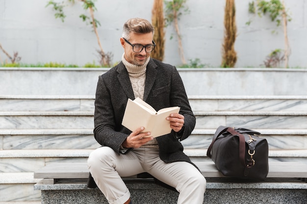 Handsome man wearing jacket reading a book while sitting outdoors