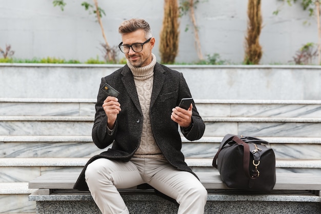 Handsome man wearing jacket shopping online with mobile phone and credit card while sitting outdoors