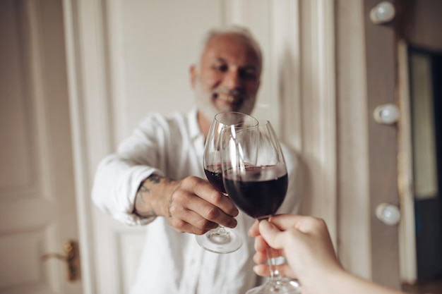 Handsome man in white outfit clinking glass with friend Smiling bearded adult guy with tattoos on his hand with red wine