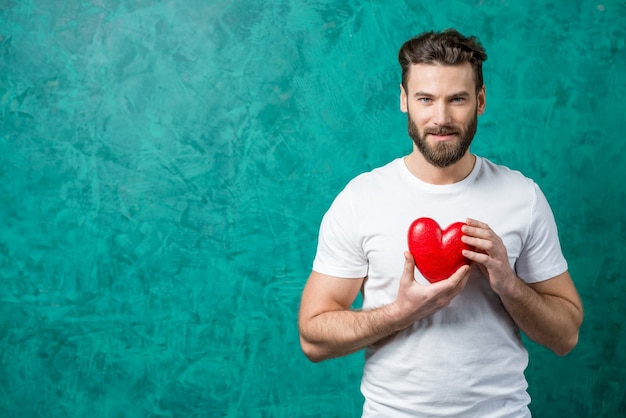 Photo handsome man in the white t-shirt holding red heart on the painted green wall background. valentine's day concept