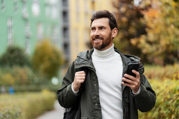 Handsome man with a beard on the city street holding a phone in his hand He has a backpack on his shoulder he looks away with a smile