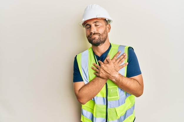 Handsome man with beard wearing safety helmet and reflective jacket smiling with hands on chest, eyes closed with grateful gesture on face. health concept.